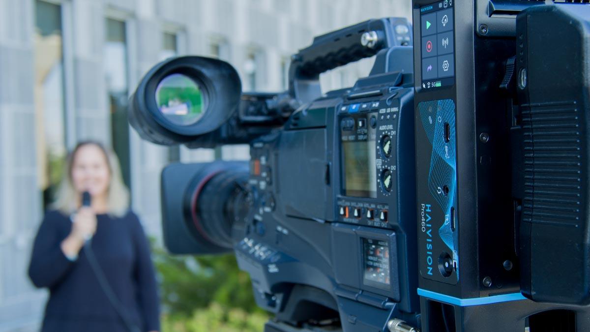 Broadcast camera with a Haivision Pro460 field encoder mounted on the back, filming a presenter on location